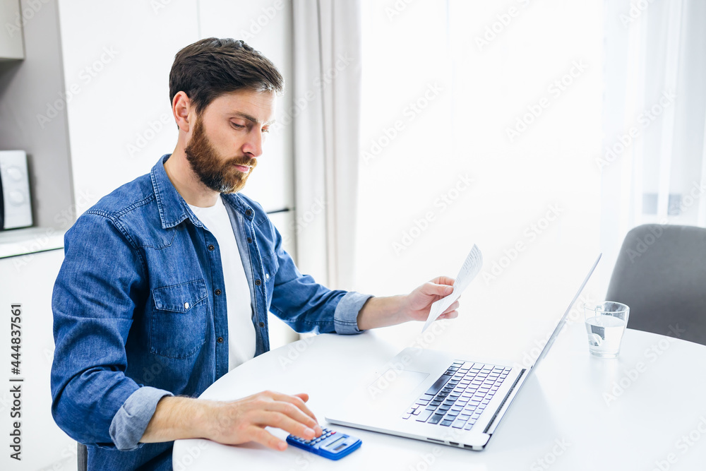 Serious man calculating domestic bills or taxes, household expenses, budget management, paperwork, checking financial documents, using laptop and calculator in modern kitchen Serious man calculating domestic bills or taxes, household expenses, budget management, paperwork, checking financial documents, using laptop and calculator in modern kitchen