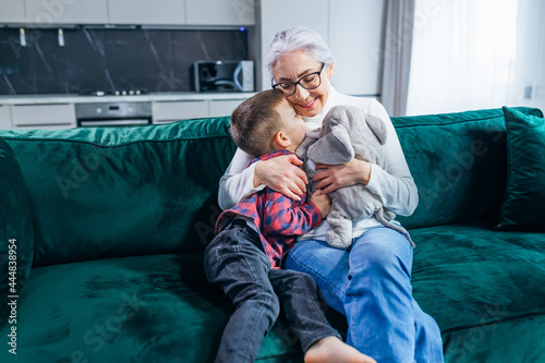 Fotografie A gray-haired grandmother with glasses plays with her grandson on the couch with soft toys and gently hugs him
