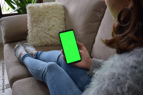 Image of a woman holding a mobile phone with a green screen while sitting on the sofa with a feeling of relaxation.
