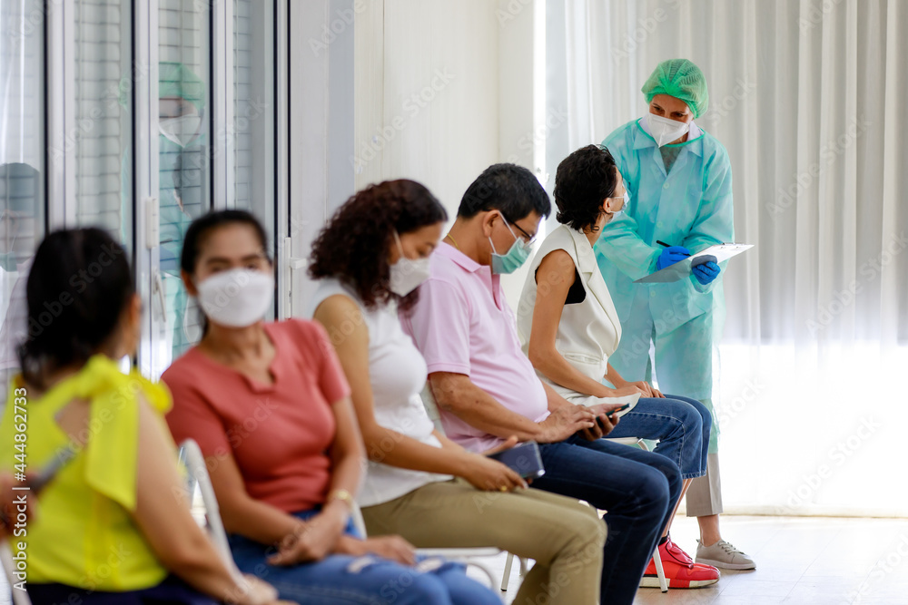 Young female citizen wears face mask in Coronavirus vaccinating queue line sitting checking her personal information from paper board in hand of full protective suit nurse who standing nearby Young female citizen wears face mask in Coronavirus vaccinating queue line sitting checking her personal information from paper board in hand of full protective suit nurse who standing nearby
