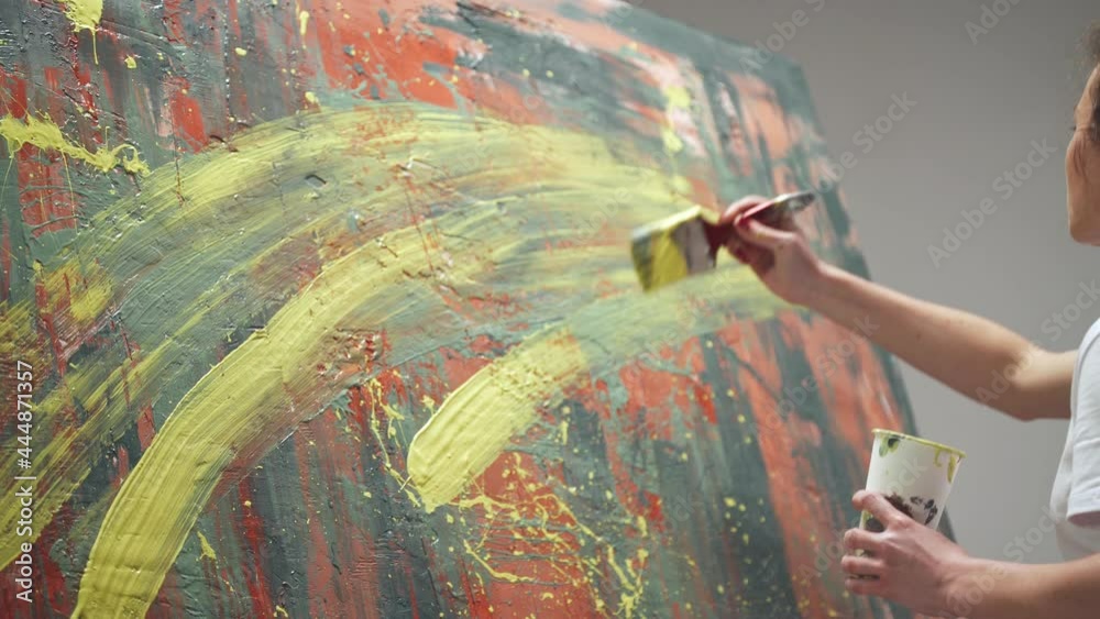 Young woman artist draws with a brush on a large canvas, a talented artist draws a color red abstraction, splashing paint on canvas, back view.