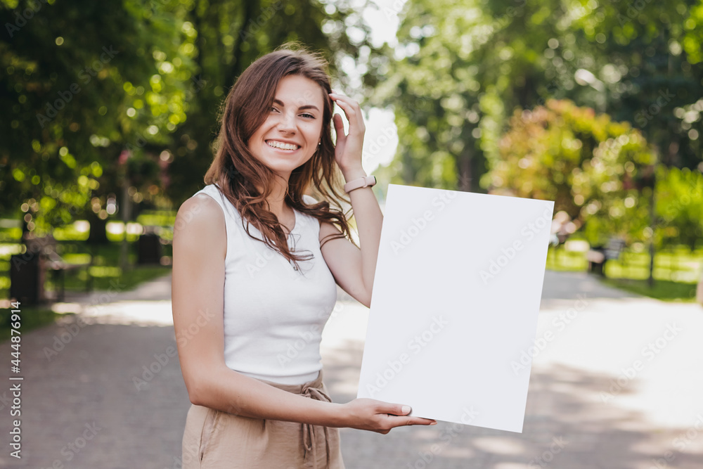 Happy caucasian young brunette woman holding a poster in her hands and smiling on the background of the park Happy caucasian young brunette woman holding a poster in her hands and smiling on the background of the park