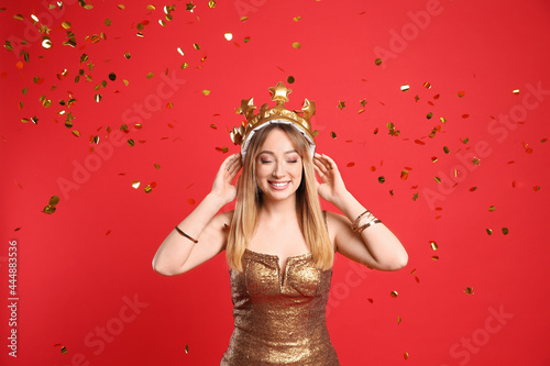 Happy young woman in party crown and confetti on red background