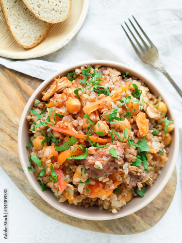 Stewed buckwheat with meat, chickpeas and vegetables, decorated with fresh herbs, served in a round bowl on a light background top view, next to a fork and two slices of bread. A delicious, hot lunch.