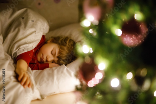 Little cute toddler girl sleeping under Christmas tree and dreaming of Santa at home, indoors. Traditional Christian festival. Happy kid child waiting for gifts on xmas. Cozy soft light © Irina Schmidt