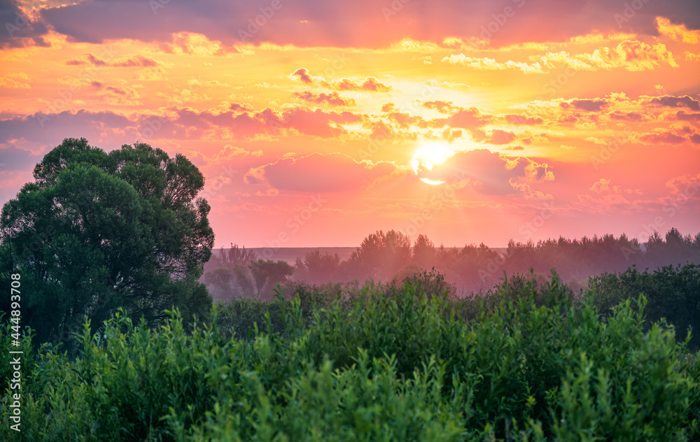Colorful sunrise in rural with a sun shining above treetops. Lush vegetation, clouds colored red, yellow, orange. Scenic view, travel, admiring, tourism, countryside beauty, sunrises and sunsets.