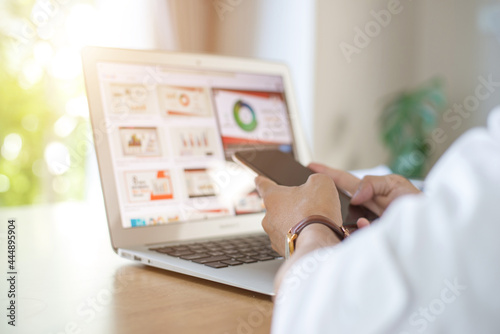 Young businessman sitting and working on a wooden desk at his home. In his hand he was holding the smart phone he was sitting and looking at the financial statements with laptop.
