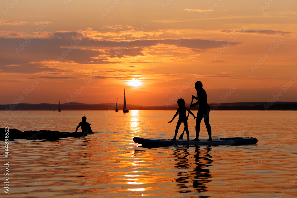 Fototapeta premium Silhouettes of a mother and daughter stand up paddle board or SUP at sunset