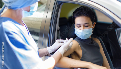 Portrait closeup shot of female wearing face mask sitting in car receiving coronavirus vaccine from doctor wears hospital uniform using syringe and needle in drive through vaccination queue