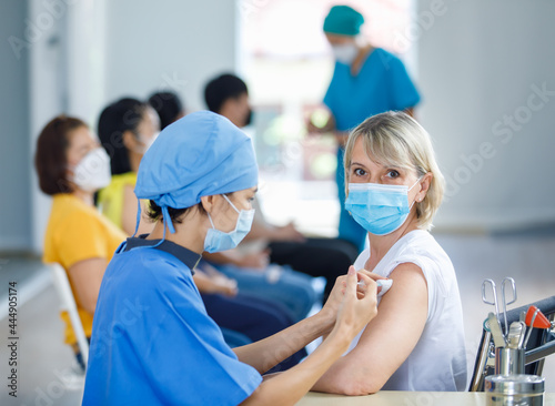 Female professional doctor wears face mask and blue hospital uniform injecting coronovirus vaccine syringe needle into Caucasian senior woman patient shoulder at clinic ward working desk