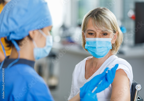 Caucasian senior patient wears face mask looking waiting to receive vaccination injection shot while doctor in blue hospital uniform preparing vaccine from glass vial dose in blurred foreground