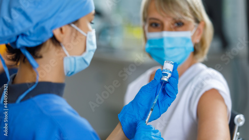 Caucasian senior patient wears face mask looking waiting to receive vaccination injection shot while doctor in blue hospital uniform preparing vaccine from glass vial dose in blurred foreground