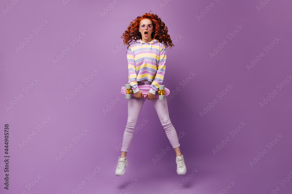 Young girl in skinny jeans and sweatshirt holds longboard and jumps on purple background Young girl in skinny jeans and sweatshirt holds longboard and jumps on purple background