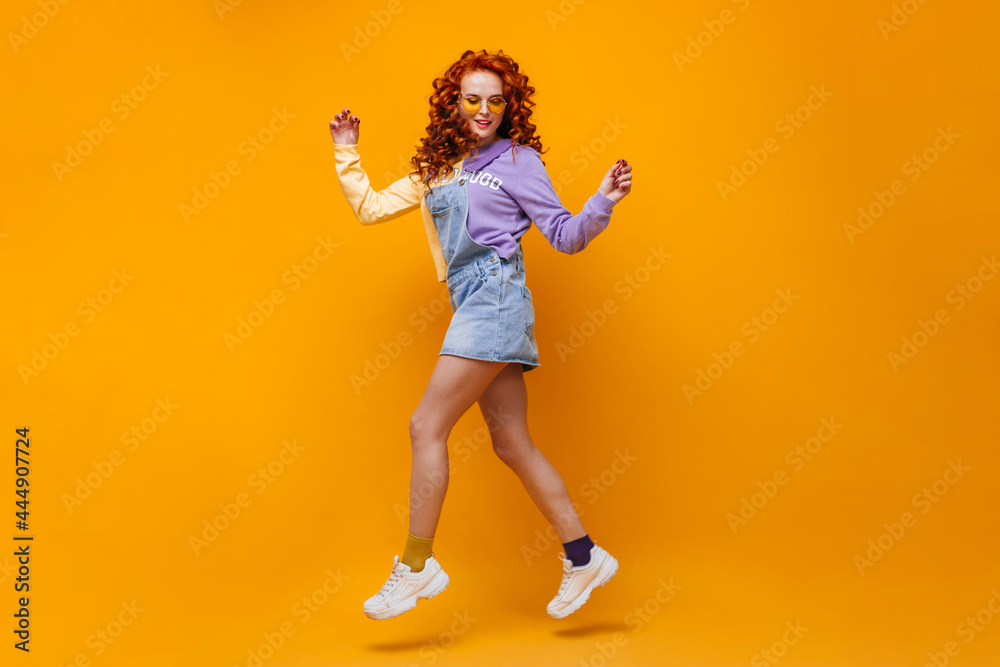 Red-haired curly girl in sunglasses, denim dress and multi-colored jumped socks on orange background