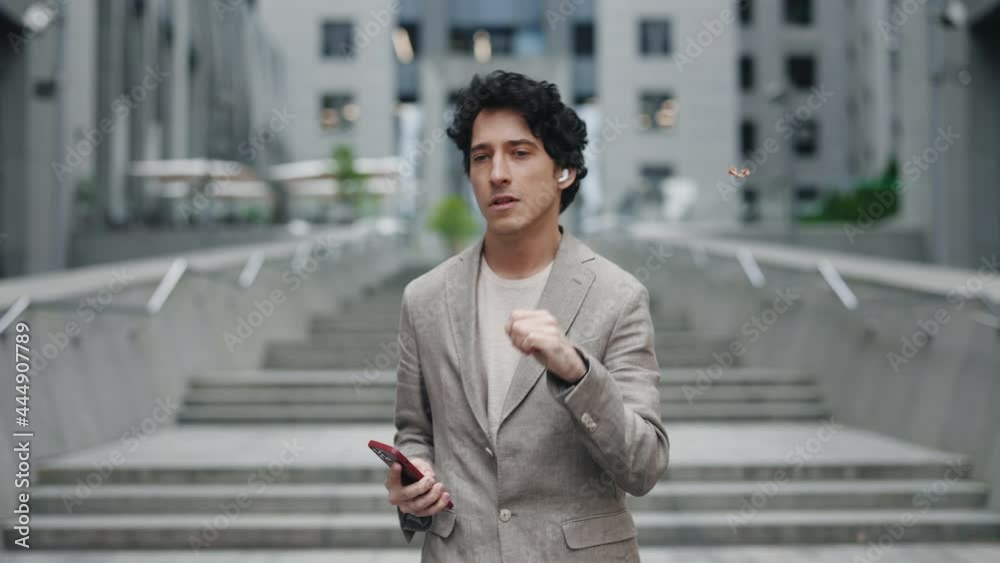 Close up of caucasian businessman wearing earphones and holding modern smartphone while walking at the street and listening music. Business people lifestyle concept. Close up of caucasian businessman wearing earphones and holding modern smartphone while walking at the street and listening music. Business people lifestyle concept.