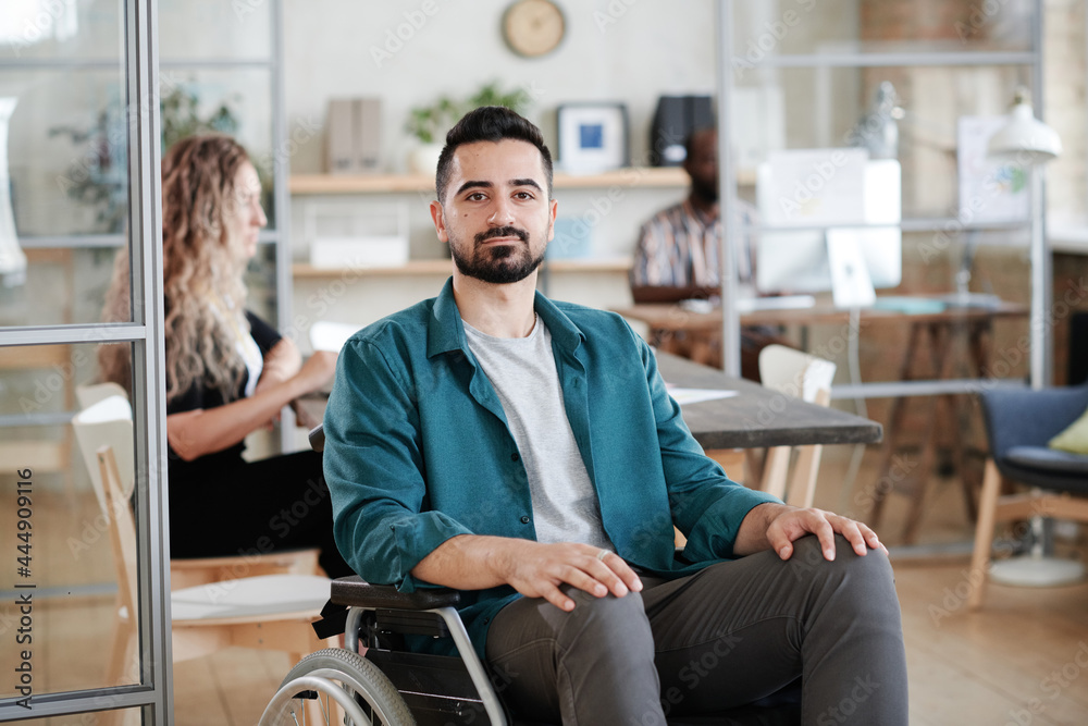 Portrait of young bearded businessman sitting on wheelchair and looking at camera while working at office Portrait of young bearded businessman sitting on wheelchair and looking at camera while working at office