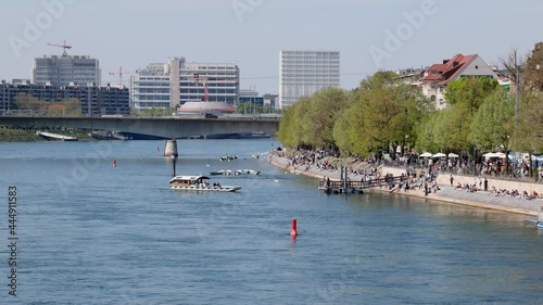 basel switzerland, april 24. 2021: view of the rhine blue passenger ferry crossing the rhine summer day many people sitting on the shore enjoying the nice weather industry in the background