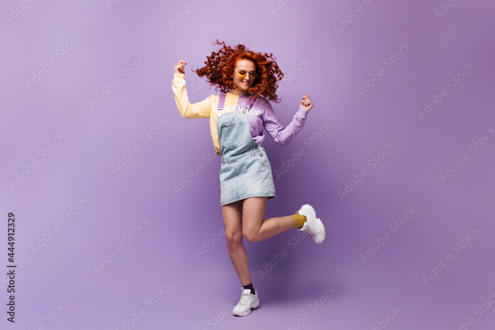 Red-haired woman in yellow glasses having fun, jumping on lilac background