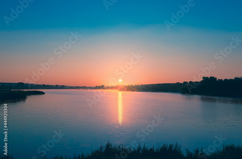 Pink Dawn: The sun rises over the blue calm water of the pond. A strip of sunlight is reflected in the blue surface of the water. Calm dawn landscape.