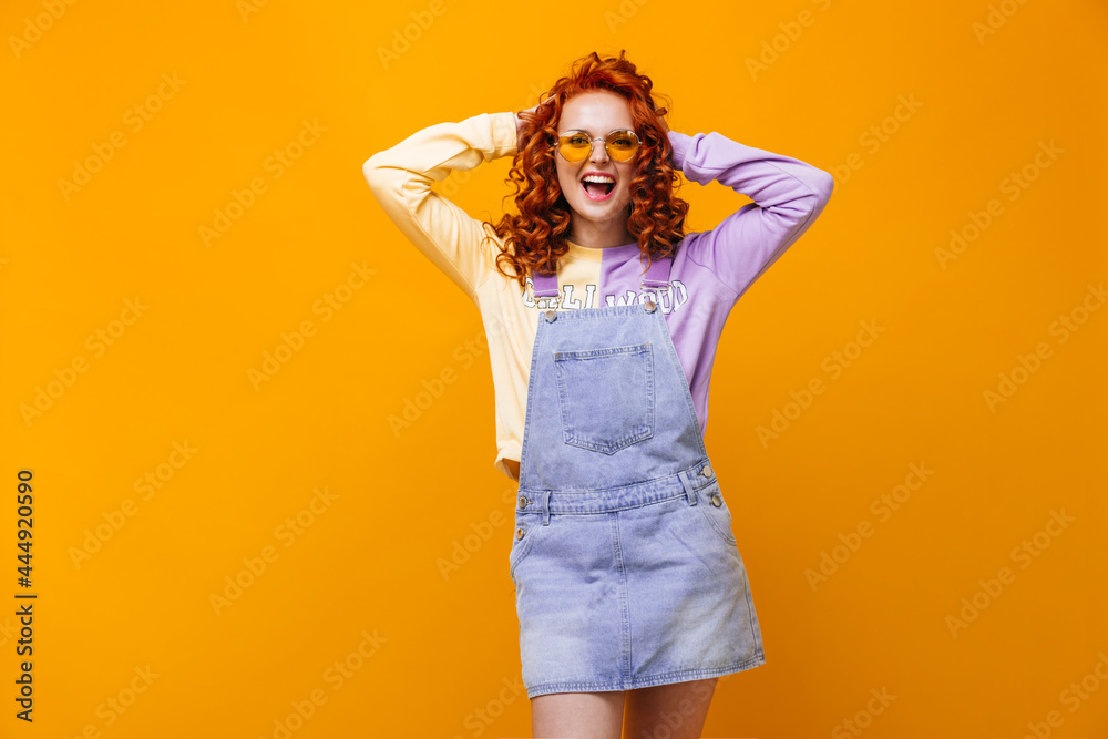 Woman with red curls tousles her hair. Girl in sundress and sweatshirt laughs on orange background Woman with red curls tousles her hair. Girl in sundress and sweatshirt laughs on orange background