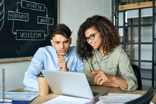 Young smiling African American mentor explaining to serious Indian coworker project strategy. Diverse startup coworkers students girl and guy talking discussing working in modern office using laptop.