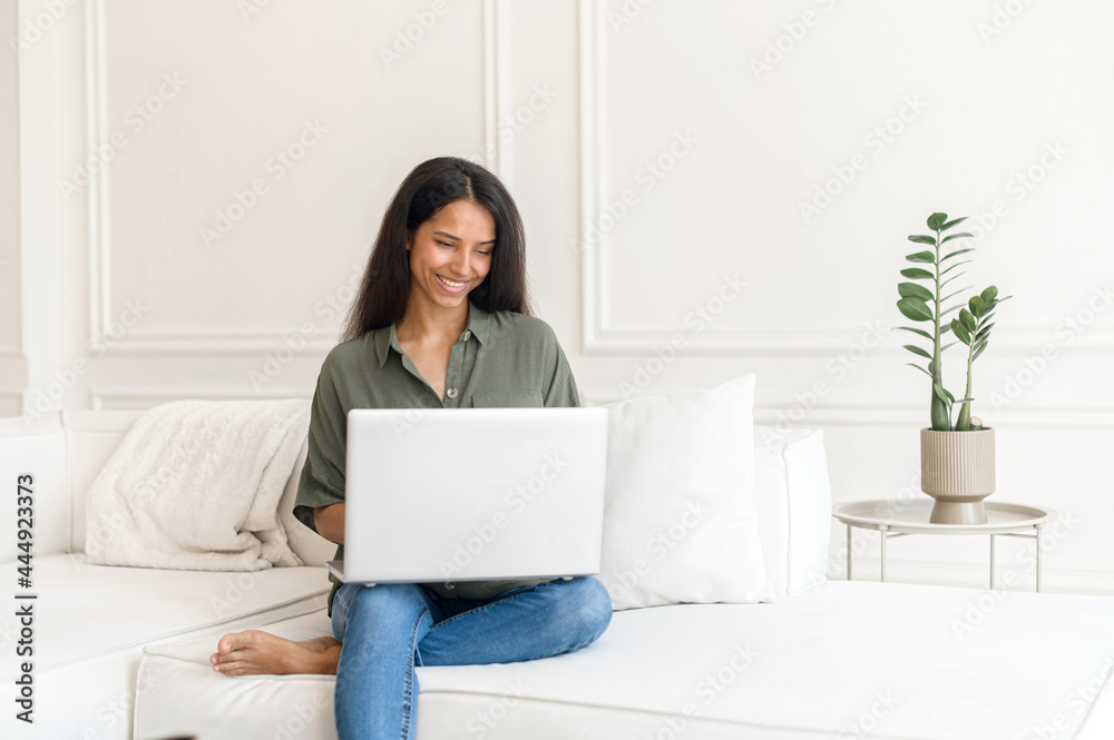 Positive intelligence indian teenage girl student sitting on the couch with laptop on the lap, studying from home, freelance woman typing email, writing article, working on project, watching video Positive intelligence indian teenage girl student sitting on the couch with laptop on the lap, studying from home, freelance woman typing email, writing article, working on project, watching video