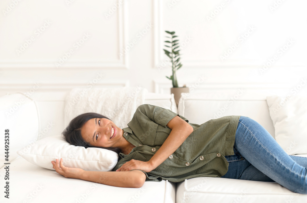 Pretty serene young asian woman is relaxing on a sofa alone. Carefree smiling indian lady lying down on the comfortable white couch and looks at the camera, resting on the pillow Pretty serene young asian woman is relaxing on a sofa alone. Carefree smiling indian lady lying down on the comfortable white couch and looks at the camera, resting on the pillow