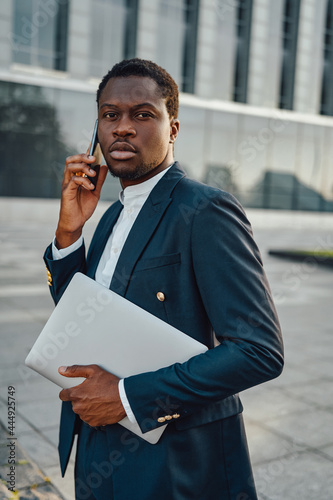 Serious black businessman with laptop talking phone outside