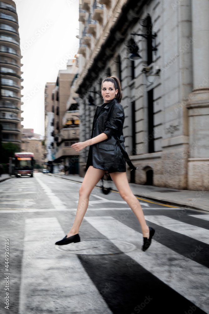 Caucasian girl dressed in black jumping in a crosswalk in the middle of the street.