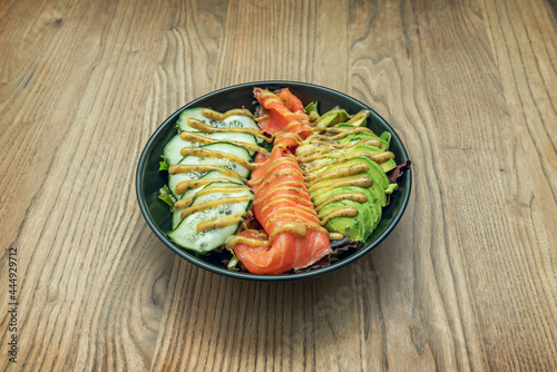 Spectacular black poke bowl with smoked salmon slices, cucumber slices, half rolled avocado, lettuce sprouts and sauce with dill on an oak table