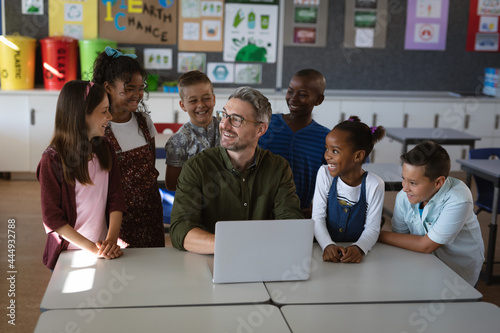 Caucasian male teacher and group of diverse students smiling while using laptop in class at school photo