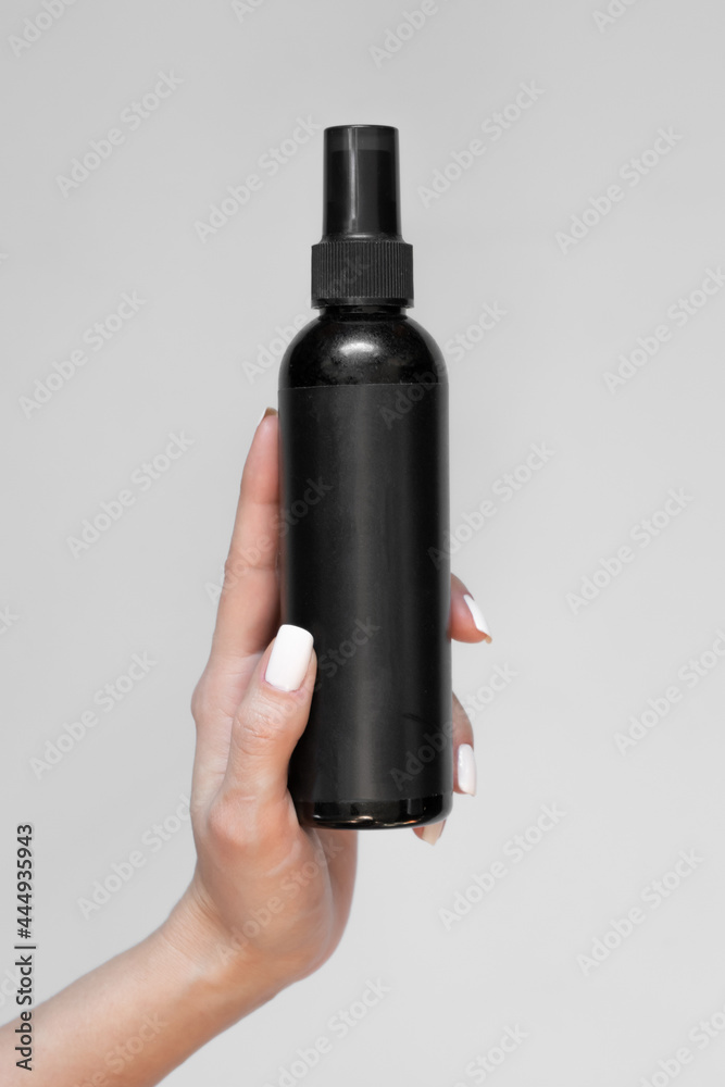 A black bottle of cosmetic spray with an empty label in a woman's hand on a light background A black bottle of cosmetic spray with an empty label in a woman's hand on a light background