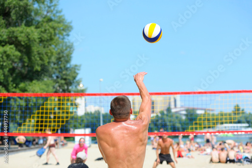 Beach volleyball. Male volley player serving the ball in front of the net, back view, blurred players