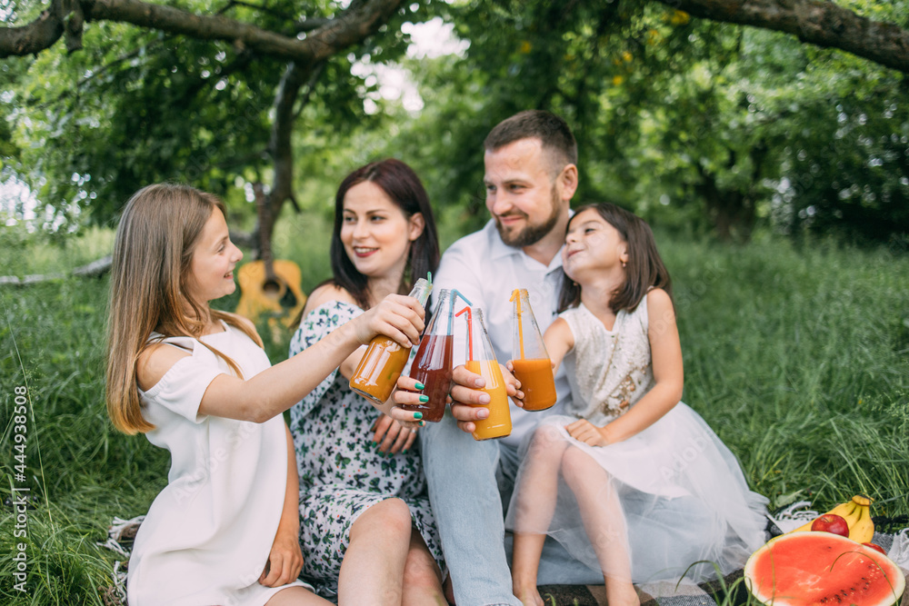 Fototapeta premium Smiling parents and their pretty daughters sitting on soft plaid outdoors and toasting with bottles of fresh juice. Family on picnic. Enjoyment concept.