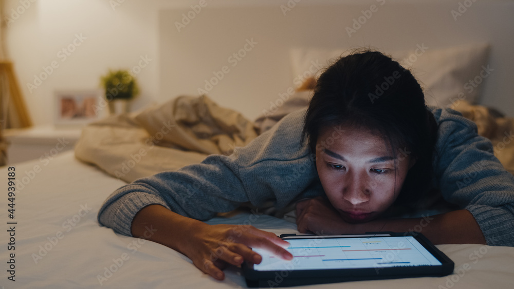 Freelance Asia women casual wear using laptop hard work on bed in bedroom at house night. Working from home, remotely work, self isolation, social distancing, quarantine for coronavirus prevention.