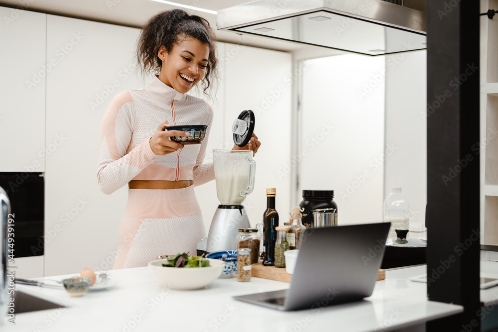 Black woman laughing and using laptop while cooking with blender Black woman laughing and using laptop while cooking with blender