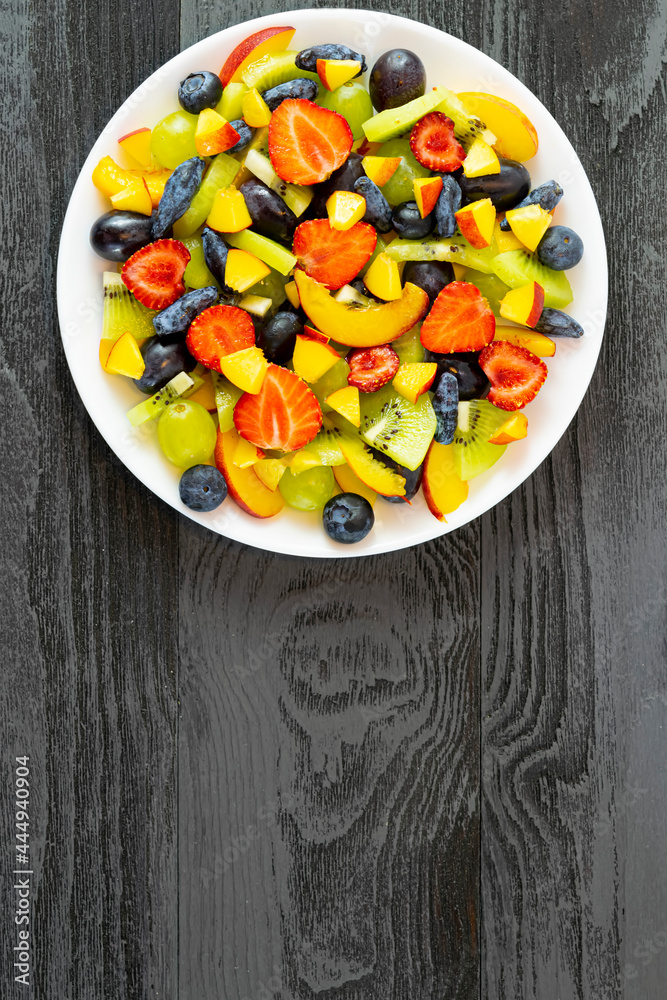 Fruit salad in a white plate, bowl in a white plate made of fresh fruits from grapes, nectarine, kiwi and strawberries, blueberries, honeysuckle on a dark wooden background.Top view, copy of the space