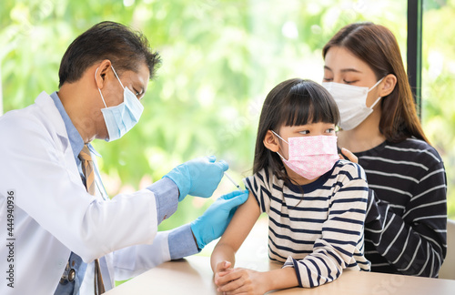 Asian senior doctor wearing gloves and isolation mask is making a COVID-19 vaccination in the shoulder of child patient with her mother at hospital.