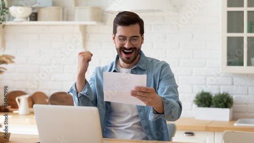 Young 35s overjoyed man sit at table hold paper sheet, read good news in letter, celebrate success about job promotion, career advance looking very happy. Great opportunity got, growth and win concept