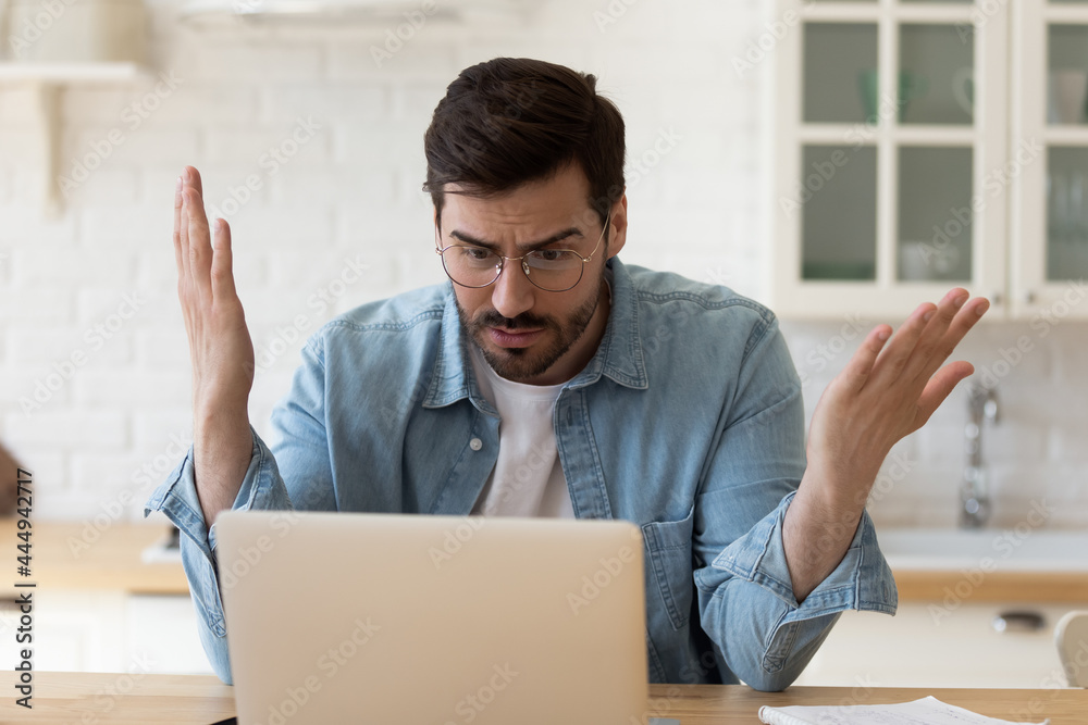 Irritated man in glasses sit at table staring at laptop screen spread arms feels angry and confused having problem with broken computer, reading bad news in email, lost information, app malfunction Irritated man in glasses sit at table staring at laptop screen spread arms feels angry and confused having problem with broken computer, reading bad news in email, lost information, app malfunction