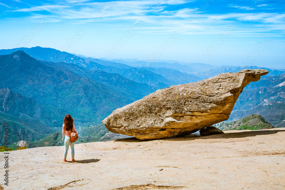 A beautiful young woman goes hiking in the mountains next to a Hanging stone in Sequoia National Park, USA. Amazing landscape from the cliff to the blue sky and mountains