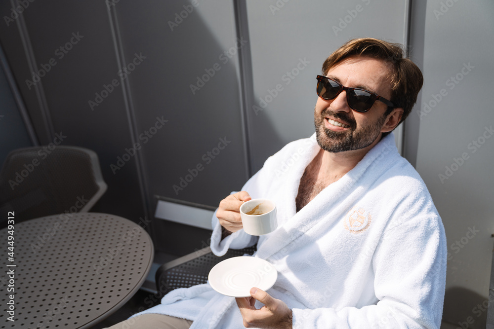 Smiling mid aged bearded man in bathrobe drinking coffee in the morning sitting on a balcony - obrazy, fototapety, plakaty 