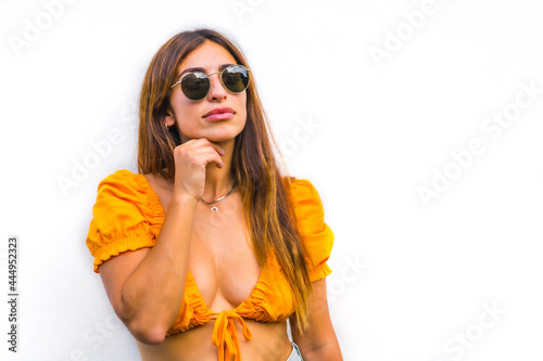 Lifestyle of a young caucasian brunette with orange t-shirt, sunglasses and short pants on a white background, with copy space and sticks. With a sweet and seductive look