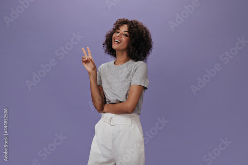 Dark-skinned woman in gray T-shirt showing sign of peace. Happy girl in white shorts smailing and posing on purple backdrop photo