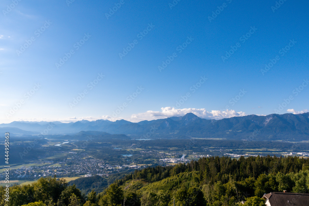 A panoramic view on the valley at the foothill of Austrian Alps. The mountains in the back are very steep and sharp. Lush pastures in front. Clear and blue sky. Serenity and calmness.