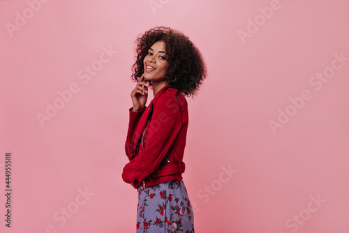 Dark-skinned woman crossed her fingers on pink background. Pretty girl in red jacket and floral dress posing on isolated backdrop