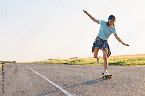 Full length portrait of happy young adult female skateboarding outdoor on asphalt road alone, raised arms, wearing casual style clothing, expressing positive emotions.
