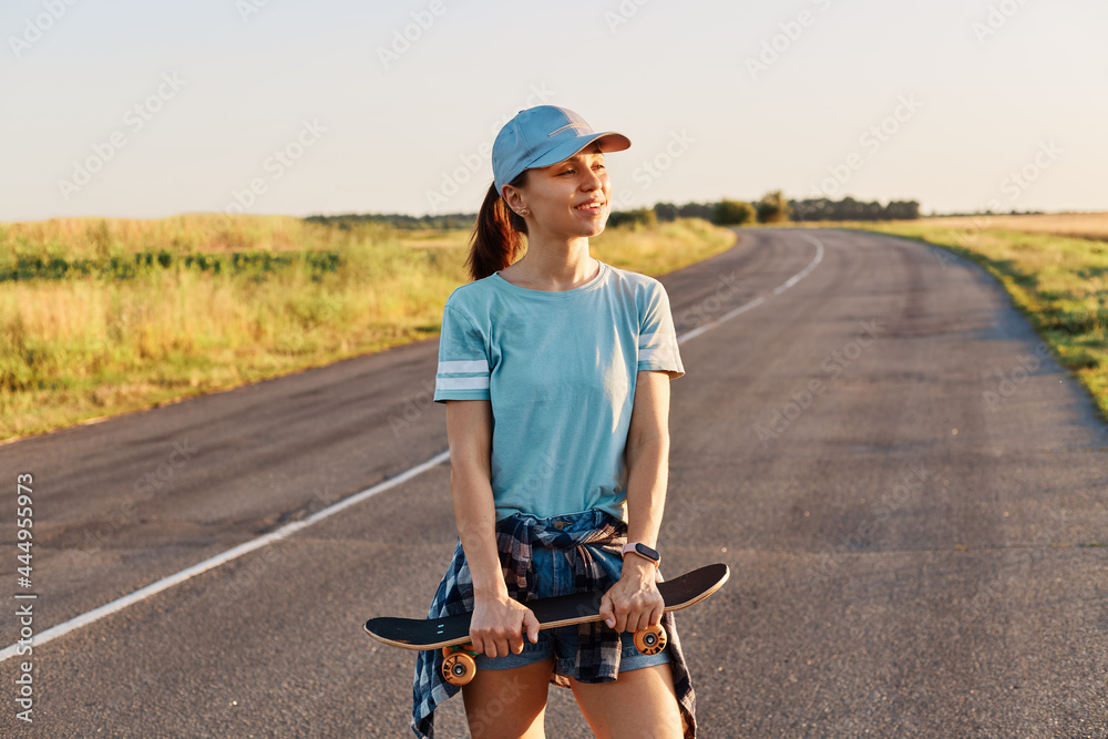 Beautiful female wearing blue t shirt and visor cap standing with skateboard in hands and looking away with toothy smile and positive emotions, active lifestyle.