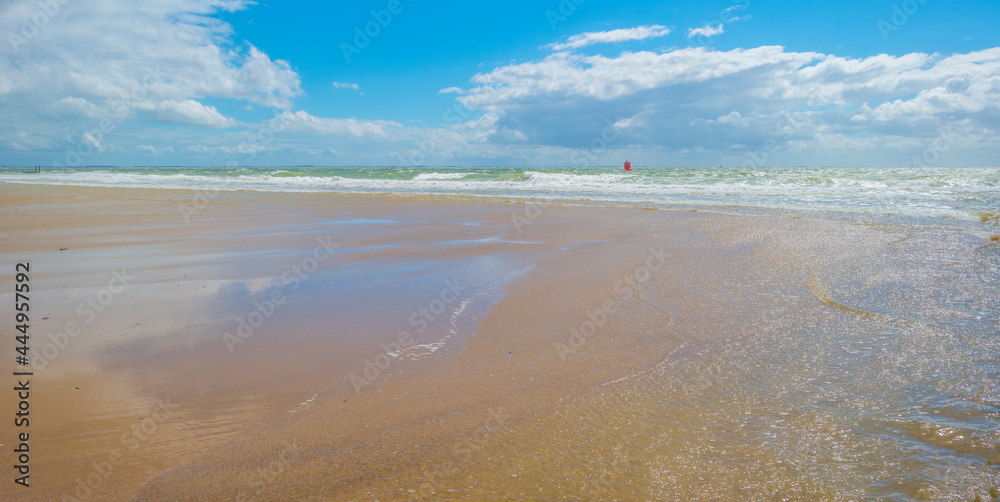 Sunlit waves on the yellow sand of a sunny beach along the North Sea illuminated by the light of a colorful sun and a blue cloudy sky in summer, Walcheren, Zeeland, the Netherlands, July, 2021
