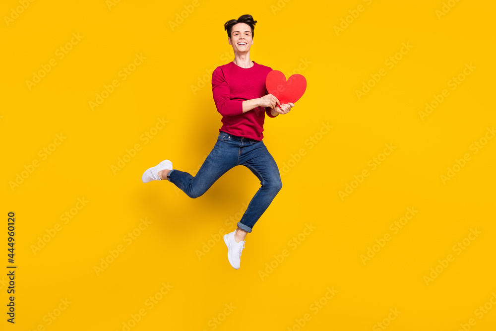 Full size photo of funny brunet young guy jump hold heart wear red sweater jeans isolated on yellow color background Full size photo of funny brunet young guy jump hold heart wear red sweater jeans isolated on yellow color background